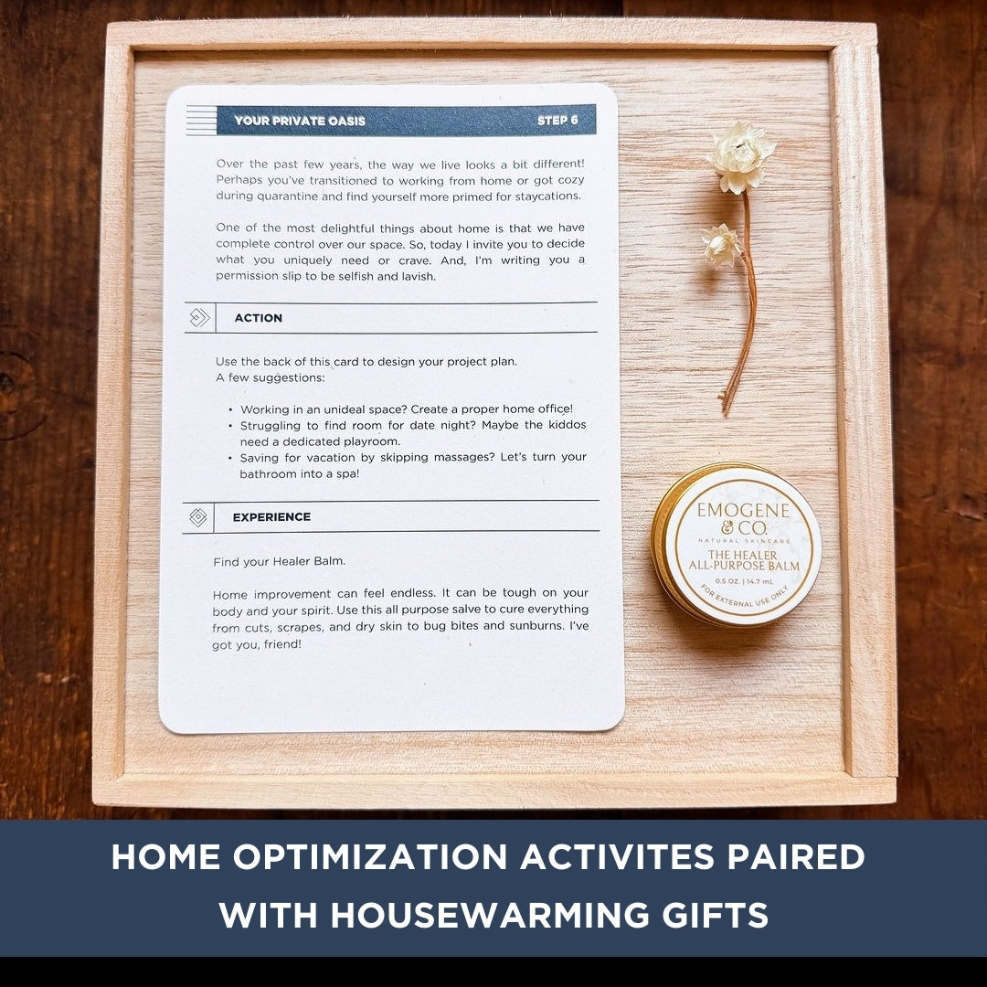 The Housewarming Box by The Box Co includes a wooden tray with home optimization tips, a golden flower, and Emogene & Co. healing balm—perfect for thoughtful gift giving. Text: HOME OPTIMIZATION ACTIVITIES PAIRED WITH HOUSEWARMING GIFTS.