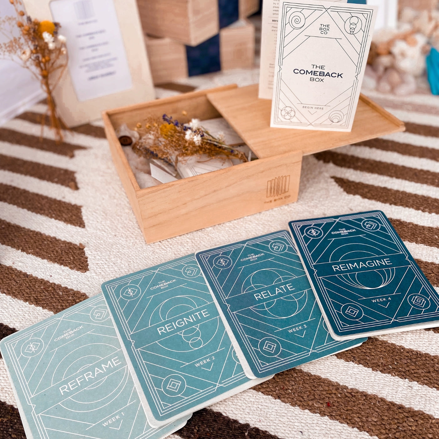 The Comeback Box: wooden box with dried flowers, 4 cards laid out with the words "reframe", "reignite", "relate", "reimagine".