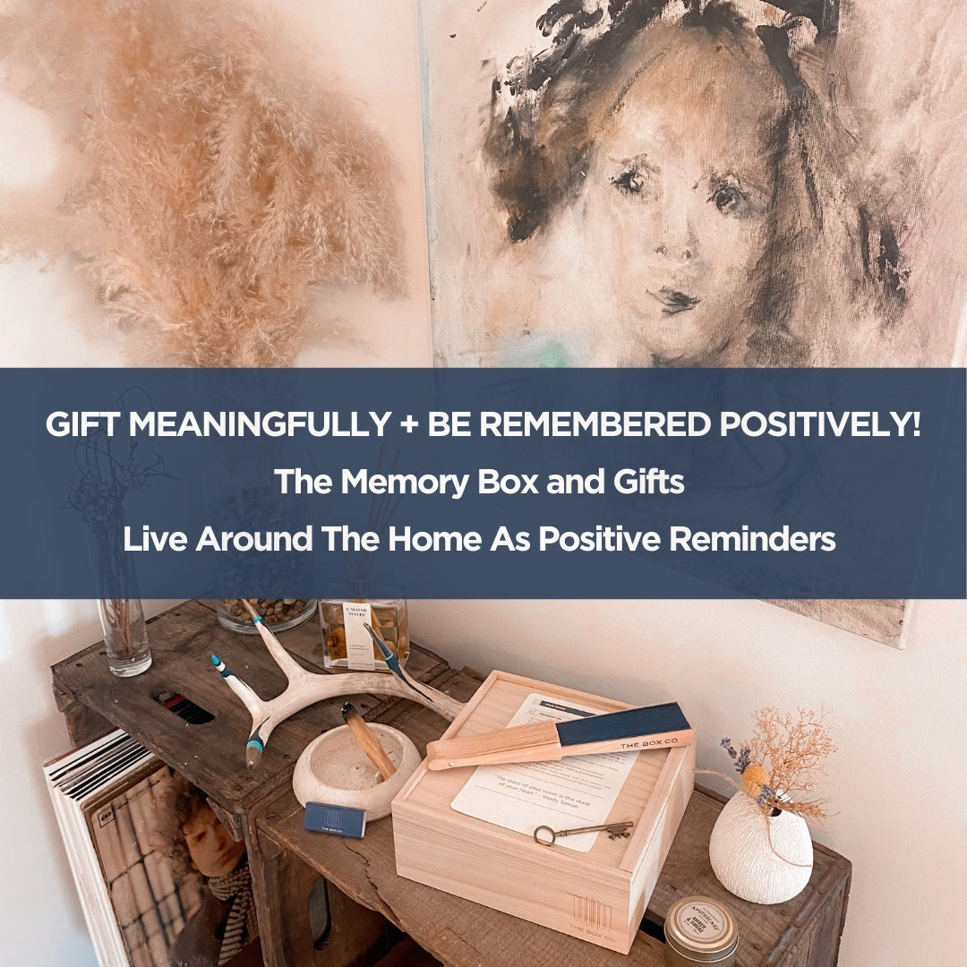 A wooden table displays The Housewarming Box by The Box Co, filled with thoughtful items for homeowners. Abstract art decorates the wall behind. Overlay: GIFT MEANINGFULLY + BE REMEMBERED POSITIVELY! Ideal for special gift occasions.
