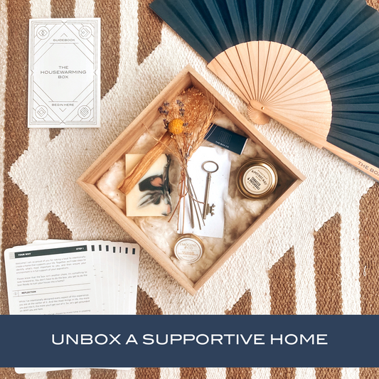 The Housewarming Box—a wooden box with dried flowers, soap, a key, candle, and more—rests on a patterned rug. A hand fan, instruction cards, and booklet are beside it. Text: “UNBOX A SUPPORTIVE HOME.” Ideal for new homeowners.