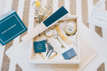 The Box Co wooden box featuring a blue hand fan, candle, soap, coaster, wildflower bouquet, and key paperweight—each symbolizing positivity—beautifully boxed with a printed list and message.