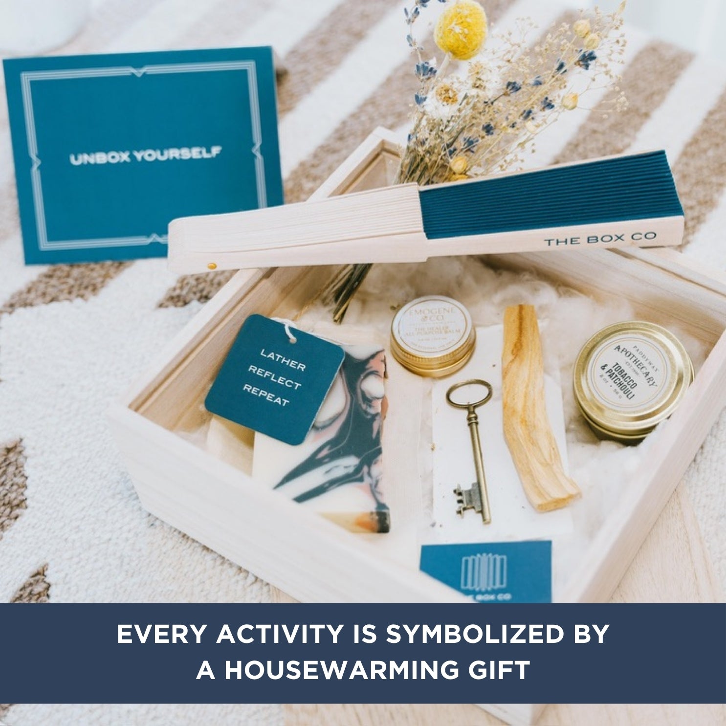 The Box Co’s Housewarming Box is a gift set for new homeowners, featuring a blue hand fan, candle, soap, coaster, wildflower bouquet, and key paperweight—each symbolizing positivity—beautifully boxed with a printed list and message.