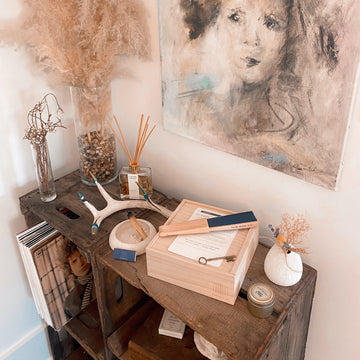 A wooden table displays wooden gift box by The Box Co, filled with thoughtfully curated items. Abstract art decorates the wall behind.