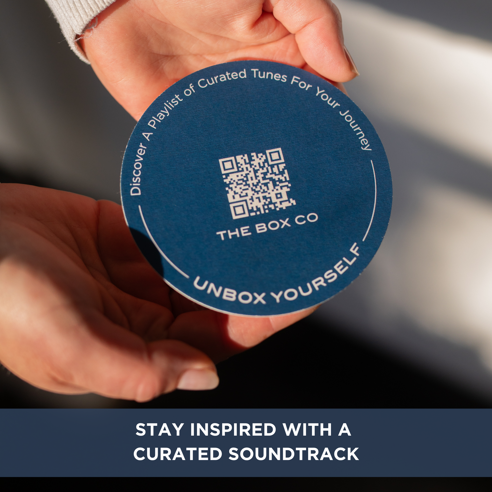 Hands holding a circular card with a QR code, The Box Co and text "Discover A Playlist of Curated Tunes For Your Journey" and "UNBOX YOURSELF". Text overlay "STAY INSPIRED WITH A CURATED SOUNDTRACK."