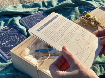 Close up of hands holding a Box Co activity card, with a wooden box and blue cards laying on a blanket on the sand.
